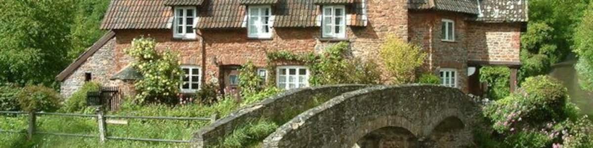 Ein Steinhaus mit einem Backsteinsschornstein, eine Steinbrücke und üppige, grüne Hügel im Hintergrund.