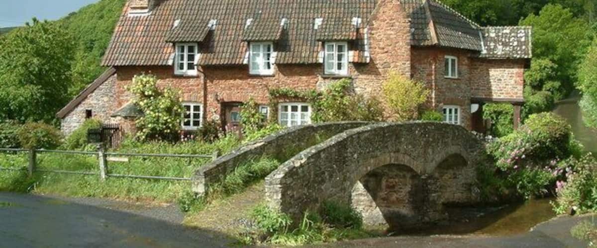 A stone house with a brick chimney, a stone bridge, and lush green hills in the background.