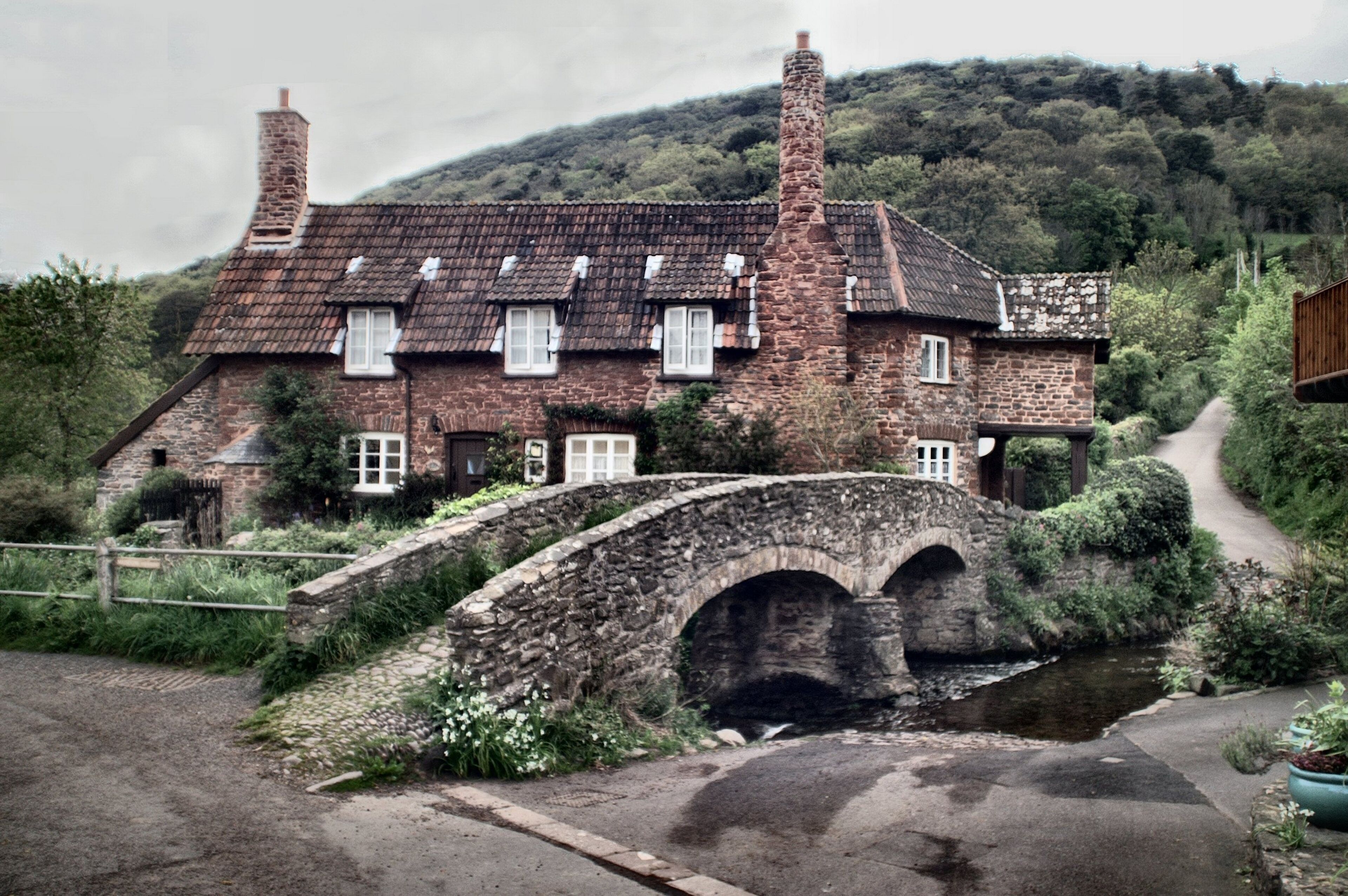 A stone house with a thatched roof, a stone bridge, and a hillside in the background.