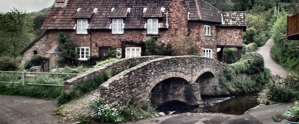 A stone house with a thatched roof, a stone bridge, and a hillside in the background.