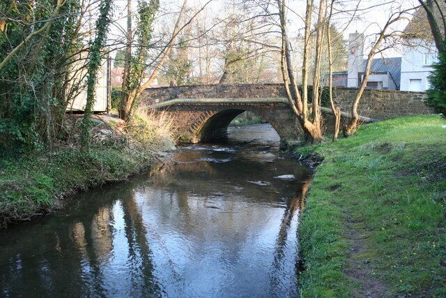 A stone bridge over a calm river, surrounded by trees and grass.