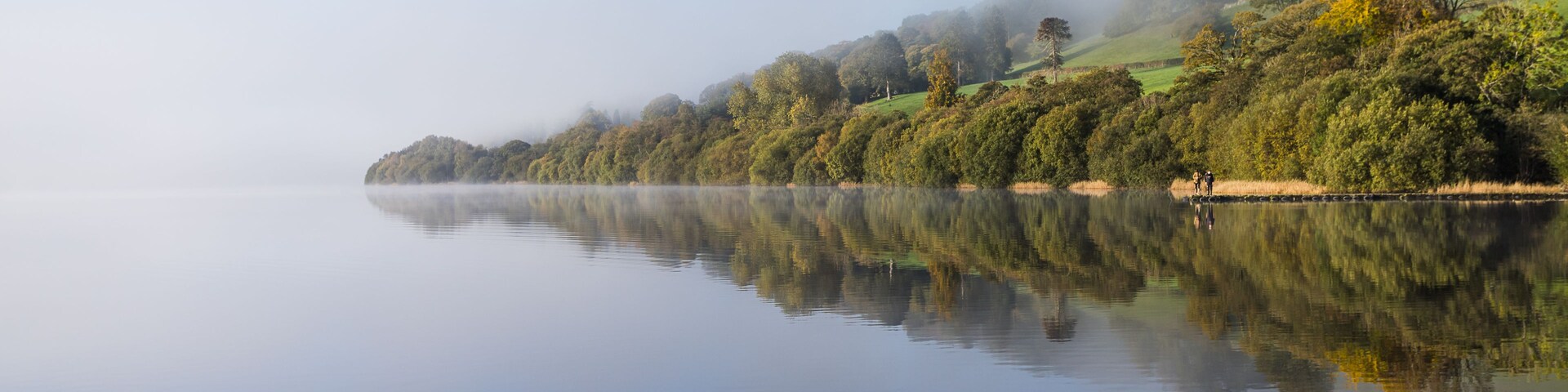 A calm lake with a misty forested hillside and a clear reflection of the trees.