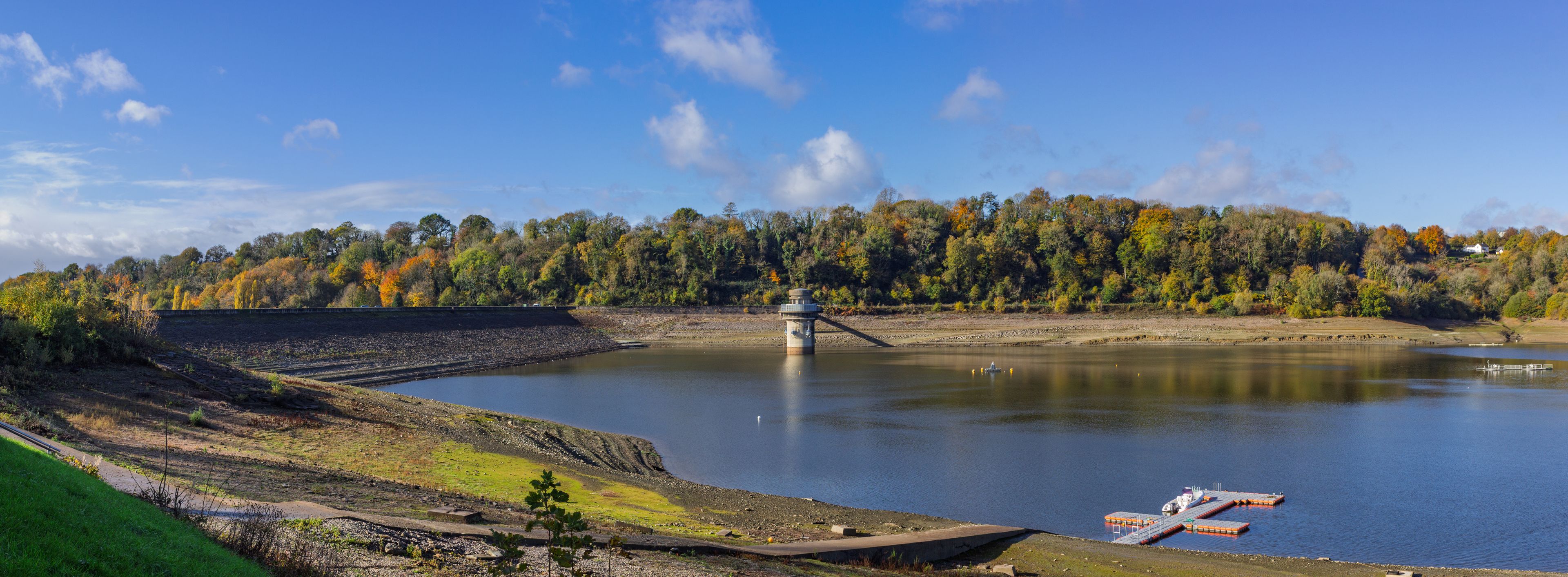 A vibrant autumnscape stretches beyond Llandegfedd lake, with its vastly reduced water level revealing the exposed embankment below the dam wall, near Pontypool, Torfaen, South Wales, UK