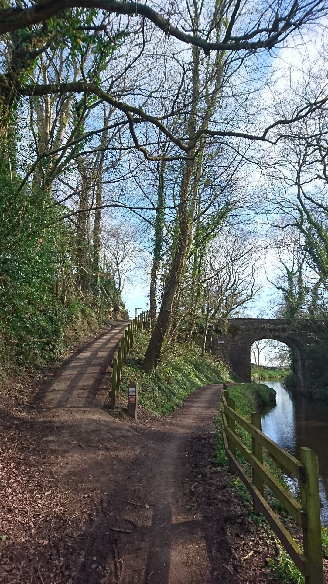 A narrow dirt path leading to a stone bridge over a river, surrounded by trees and greenery.