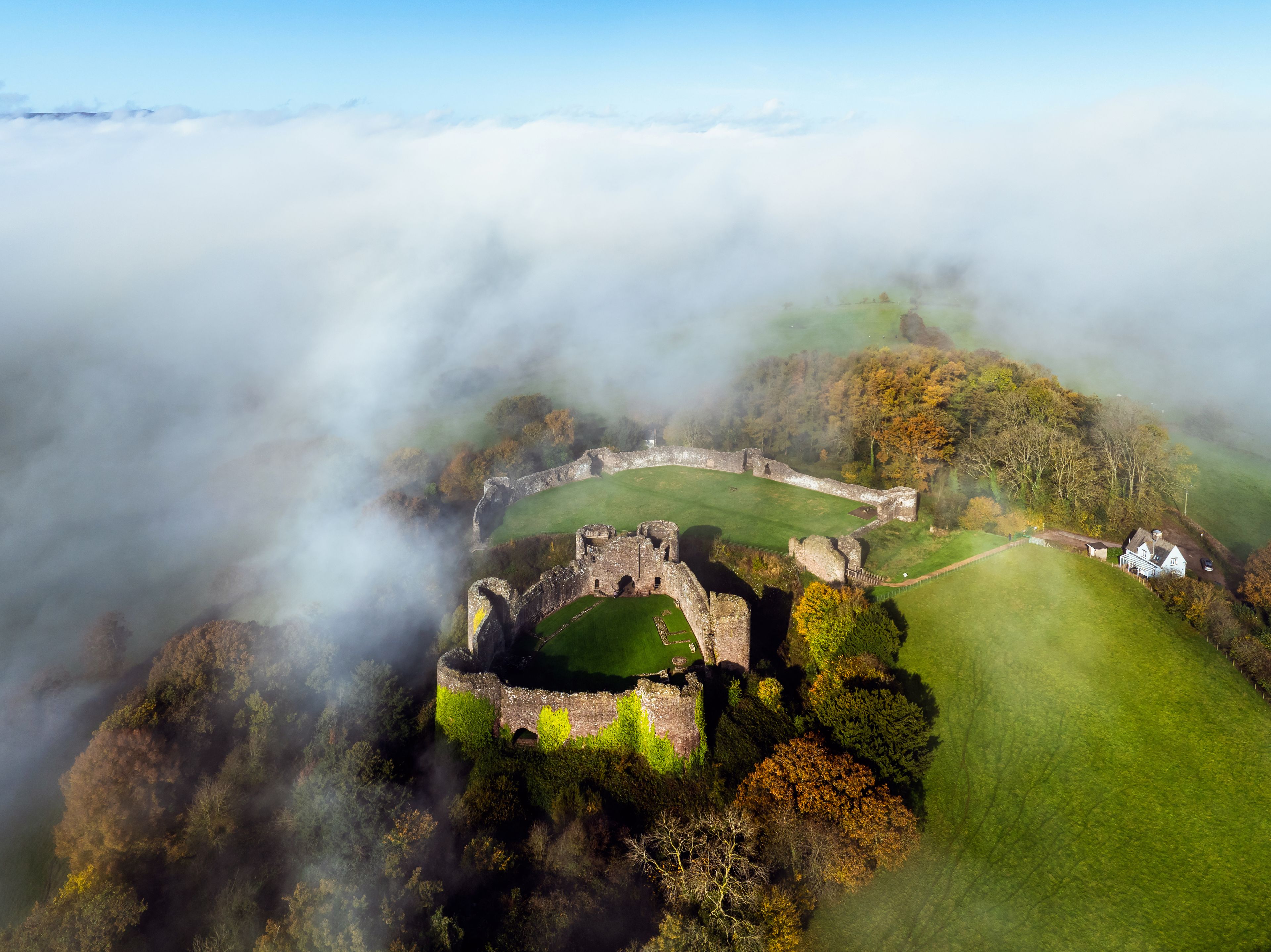 Vista aérea de un castillo histórico rodeado de niebla, con un campo verde y una casa blanca cerca.