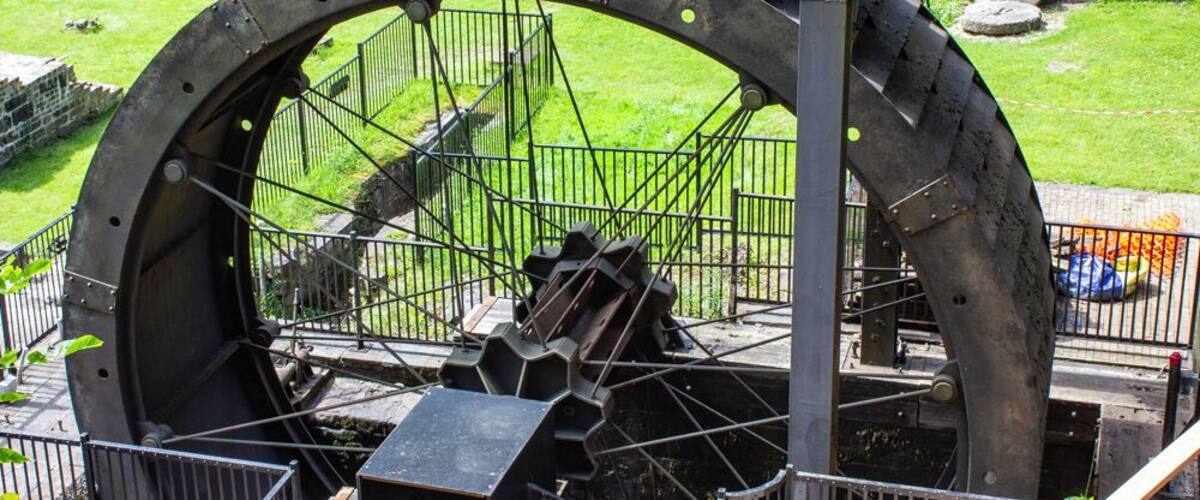 Ein großes Wasserrad mit Metallkonstruktion im Vordergrund, umgeben von einem eingezäunten Bereich mit Gras und einer Steinmauer im Hintergrund.