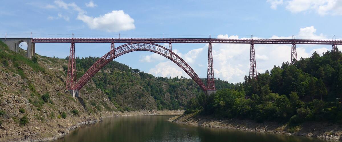 A red steel bridge over a calm river with hills on either side.
