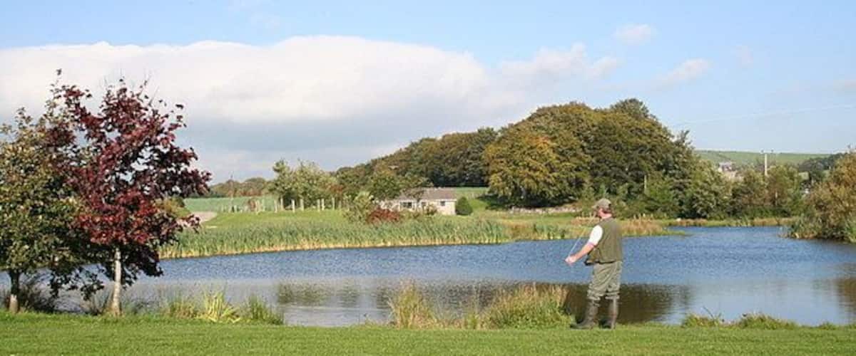 A man standing by a pond in a grassy field with trees and a house in the background.