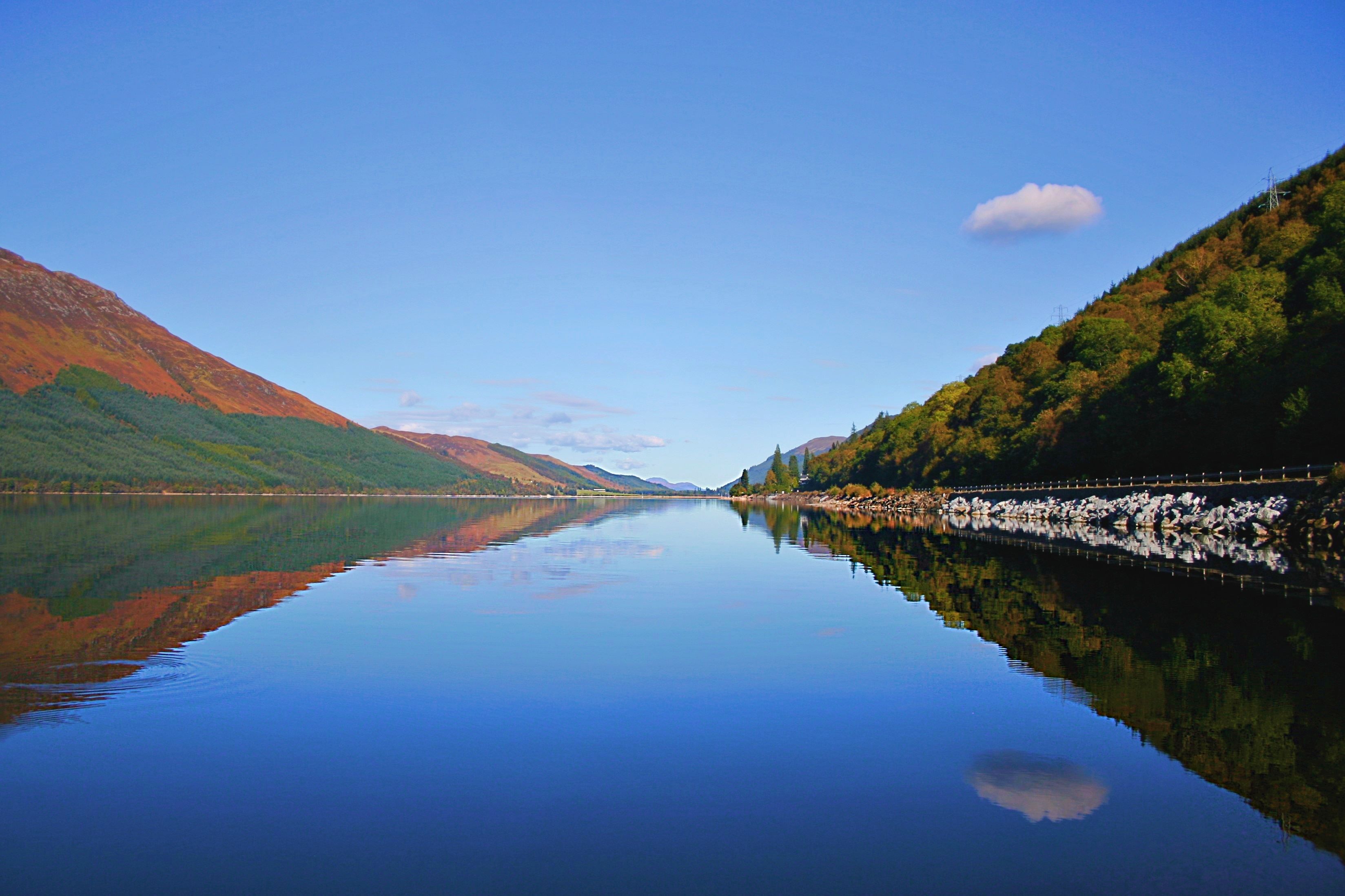 A calm river reflecting the surrounding mountains and clear sky.
