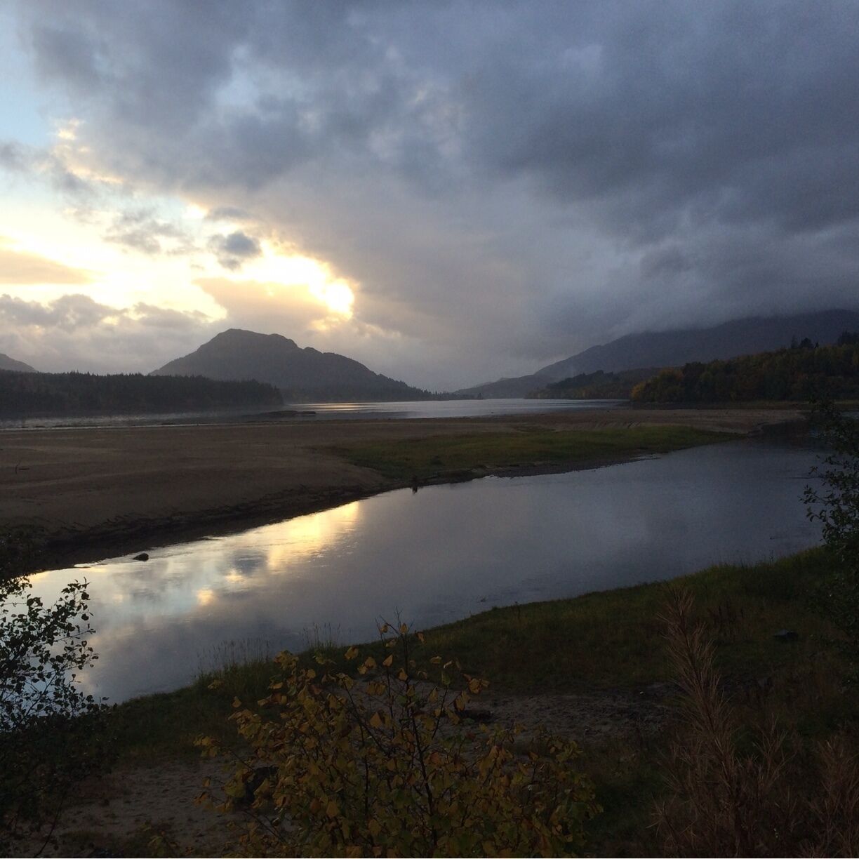 A river reflecting the sky, surrounded by grassy banks and distant mountains.