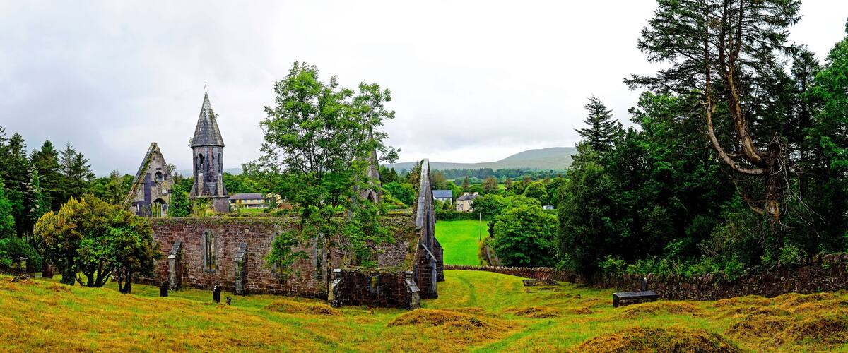 An old stone church with a tall spire, surrounded by lush greenery and a grassy field.