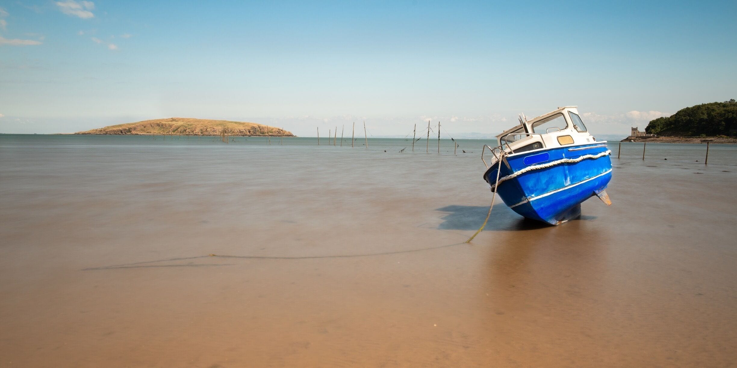 A blue boat tied to a sandy beach with a distant hill and a structure on the right.