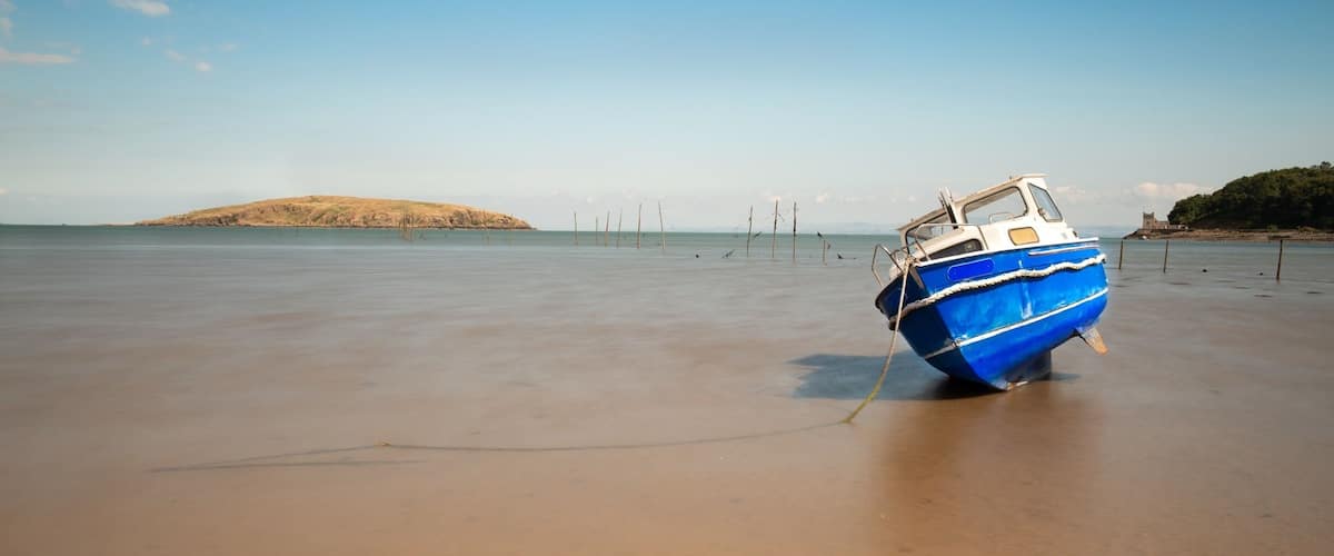 A blue boat tied to a sandy beach with a distant hill and a structure on the right.