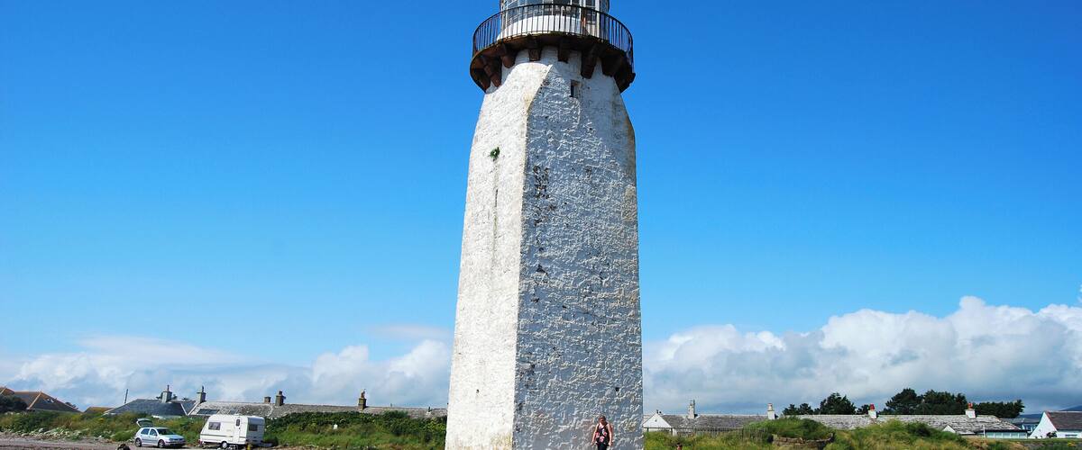 A tall, white lighthouse with a black top and balcony, surrounded by a rocky landscape and a clear blue sky.