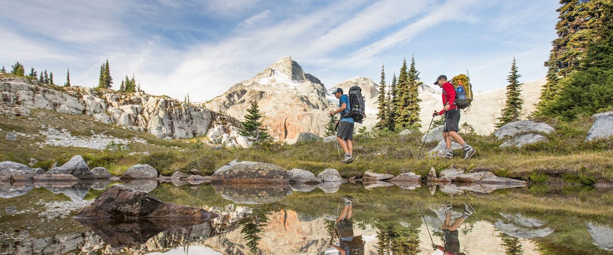 Two hikers walking on a rocky path with a mountain in the background and a reflective pond in the foreground.