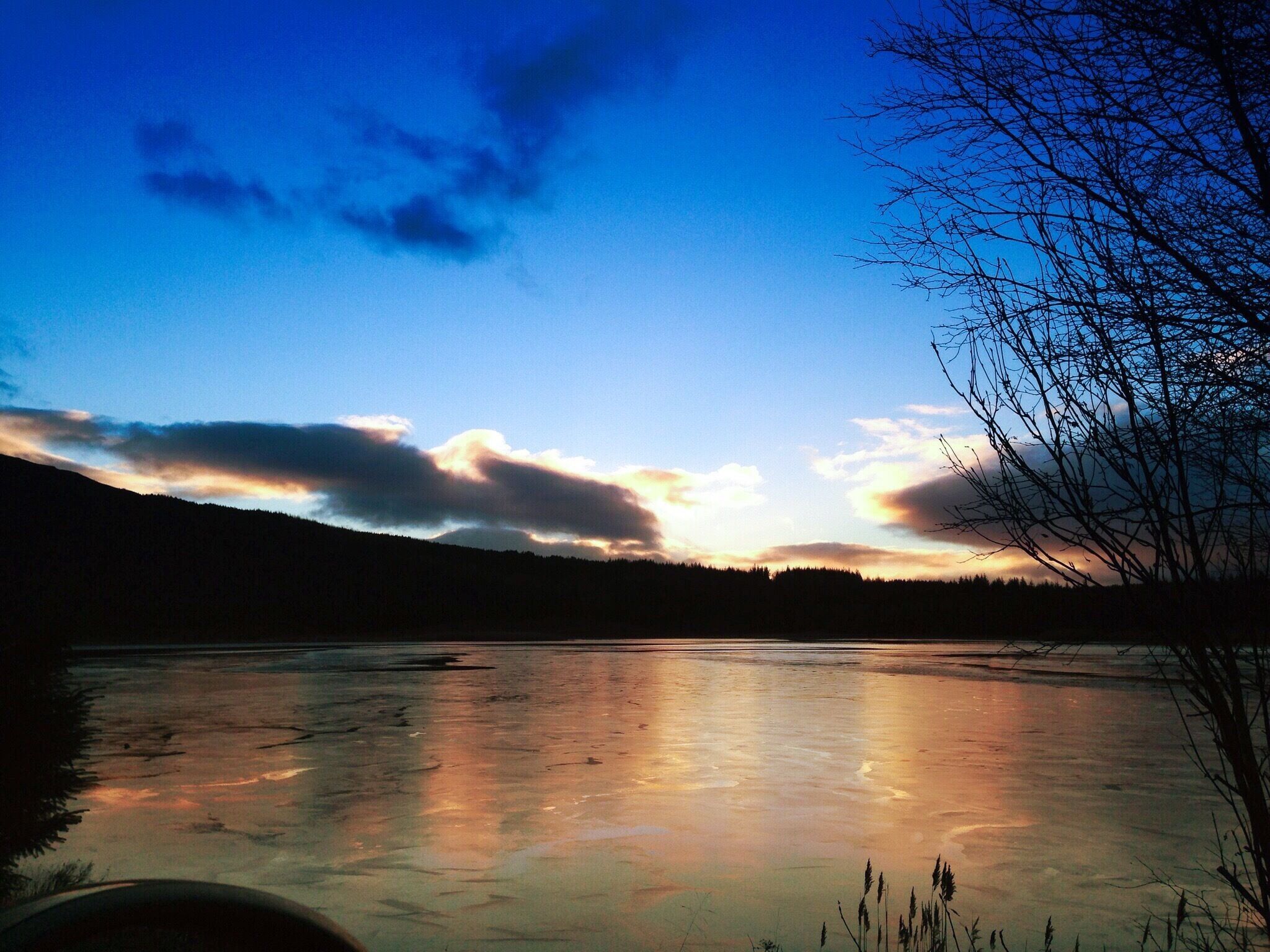 A frozen lake with reflections of the sky, surrounded by trees and hills.