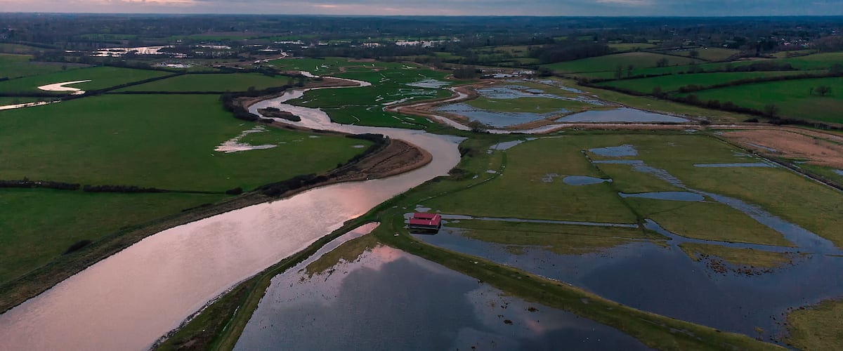 Un río serpentea por campos verdes con un edificio rojo cerca del agua.