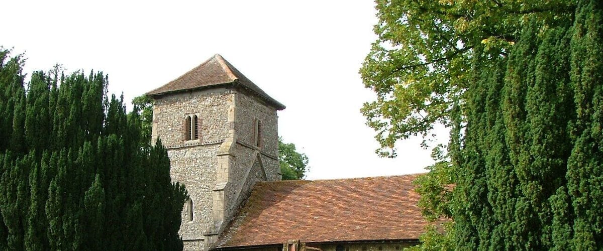 A stone church with a tower and arched entrance, surrounded by a grassy field and tall trees.