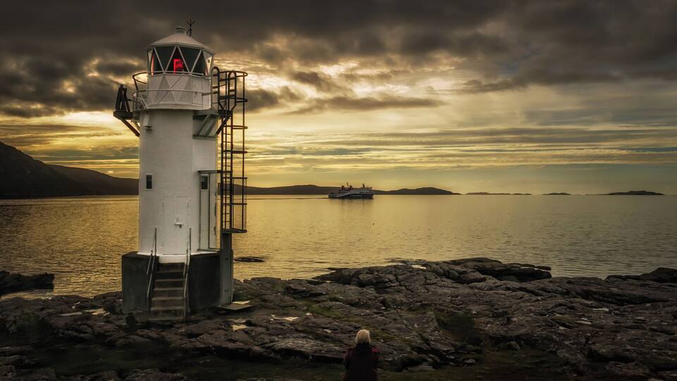 A lighthouse on rocky shore with a boat in the distance and a cloudy sky.