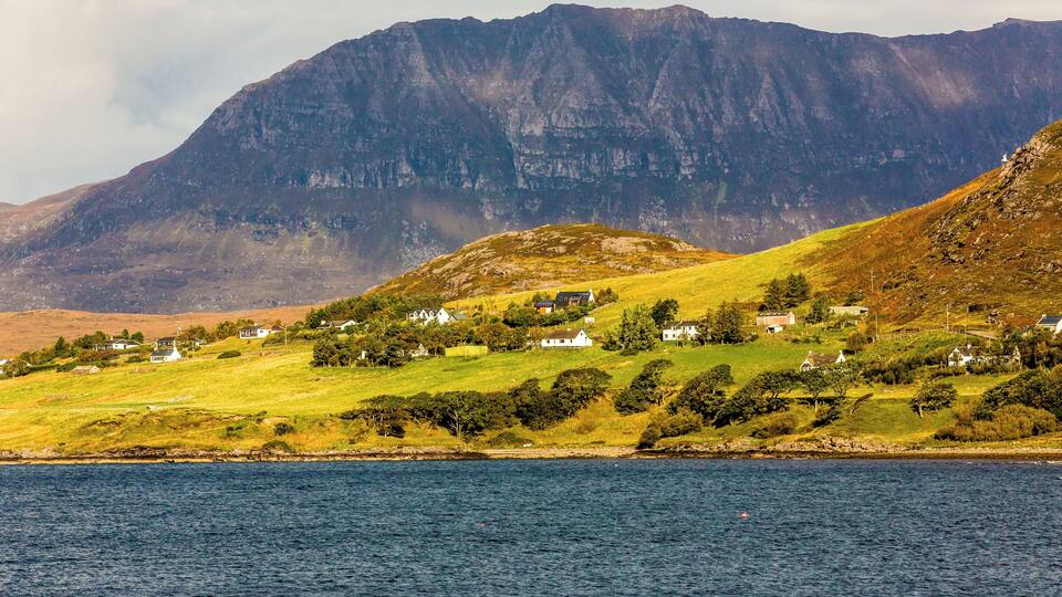 A coastal village with houses on a hillside, a mountain in the background, and a body of water in the foreground.