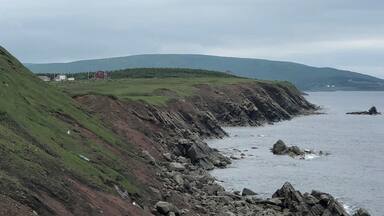 A coastal landscape with rocky cliffs, grassy slopes, and a distant view of a red building.