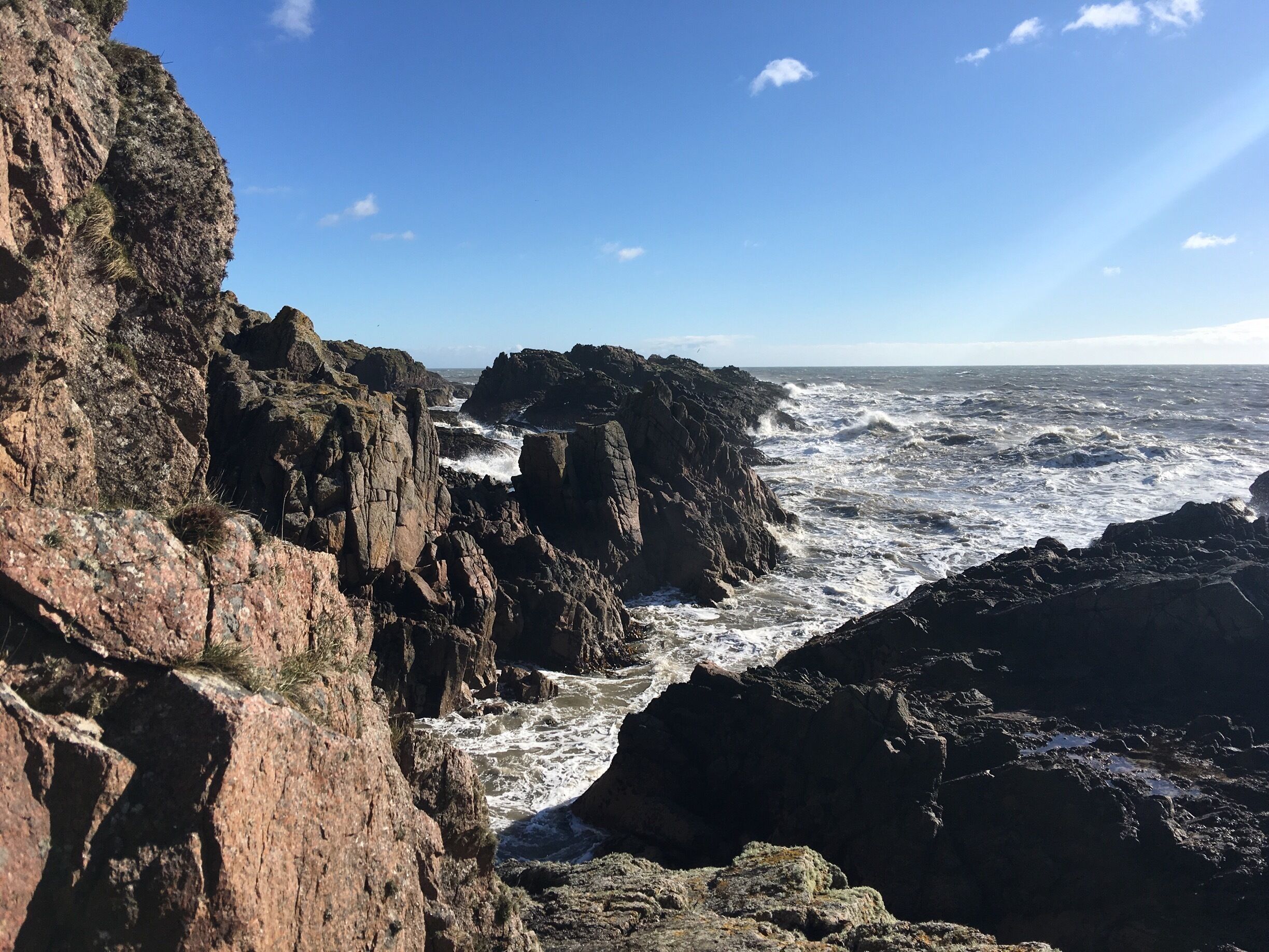 A rocky coastline with turbulent waves crashing against the cliffs under a clear sky.