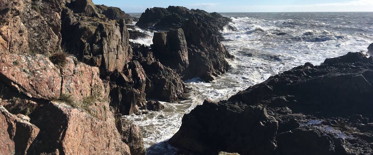 A rocky coastline with turbulent waves crashing against the cliffs under a clear sky.