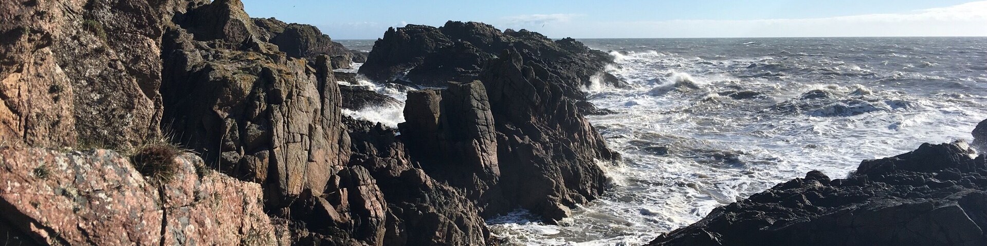 A rocky coastline with turbulent waves crashing against the cliffs under a clear sky.