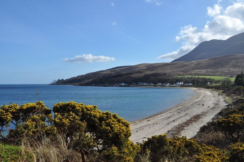 A coastal landscape with a sandy beach, a clear blue sea, and a hill with sparse vegetation.