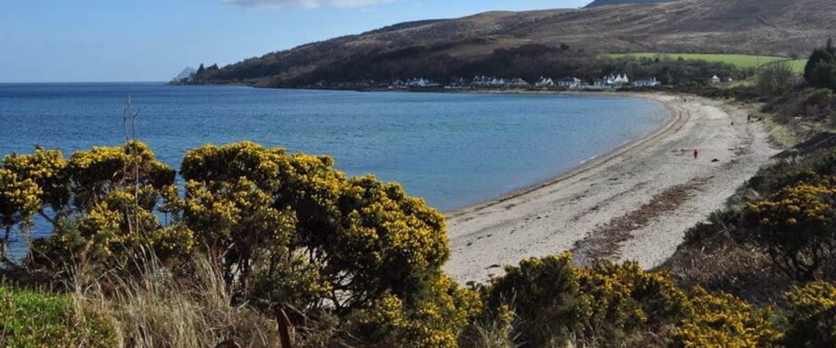 A coastal landscape with a sandy beach, a clear blue sea, and a hill with sparse vegetation.