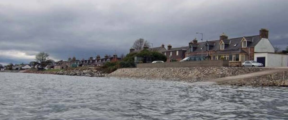 A coastal residential area with houses along the shoreline, a rocky breakwater, and overcast skies.