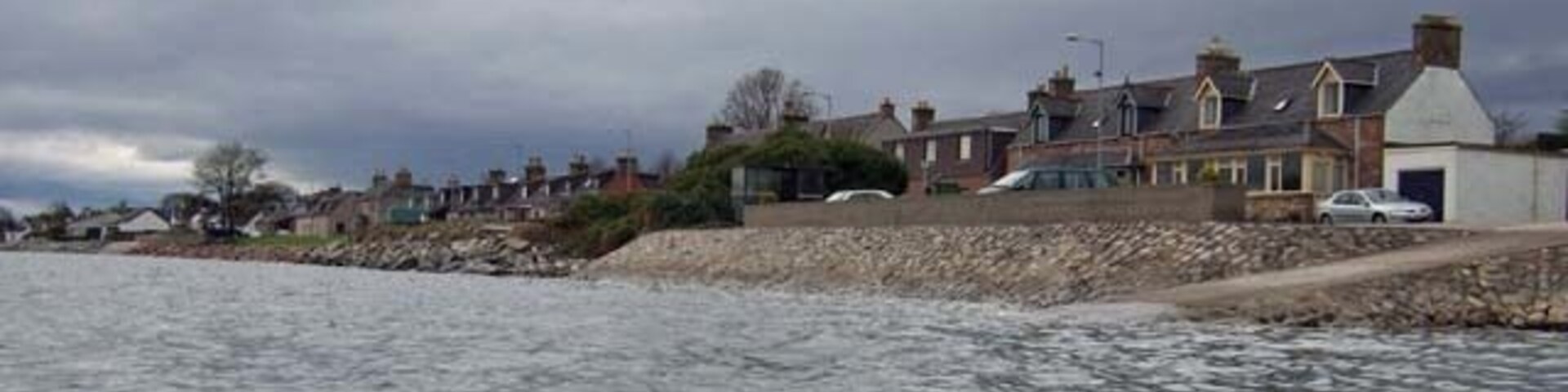 A coastal residential area with houses along the shoreline, a rocky breakwater, and overcast skies.