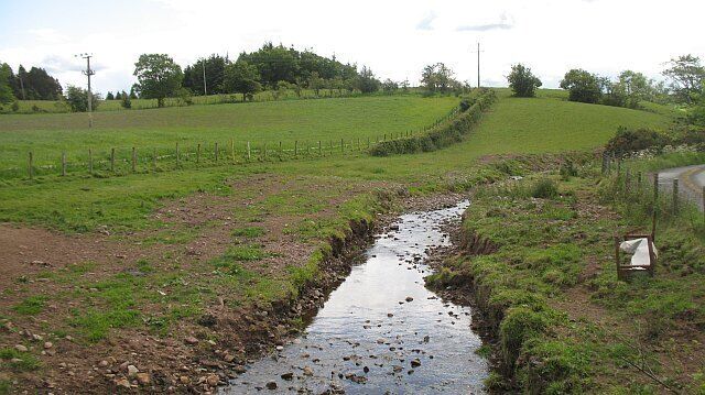 A rural landscape with a narrow stream, green fields, and a fence.