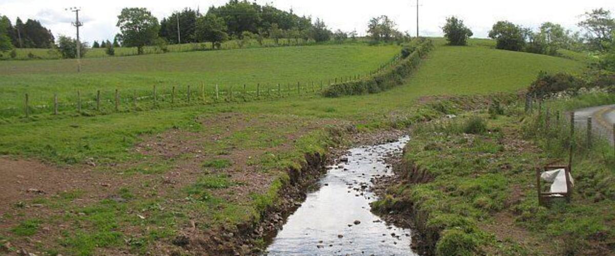 A rural landscape with a narrow stream, green fields, and a fence.