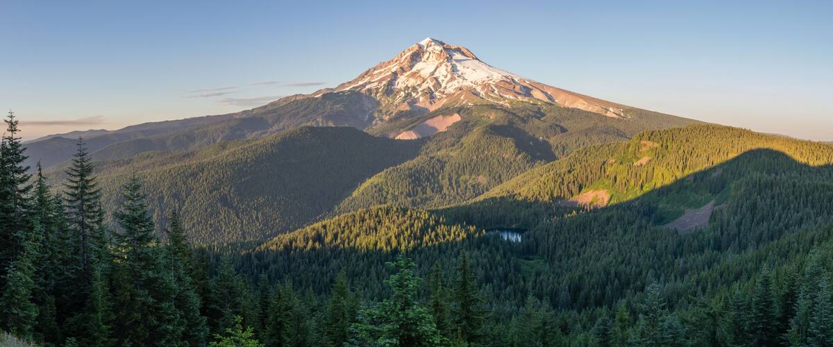 A mountain with a snow-capped peak, dense forest, and a lake at its base.