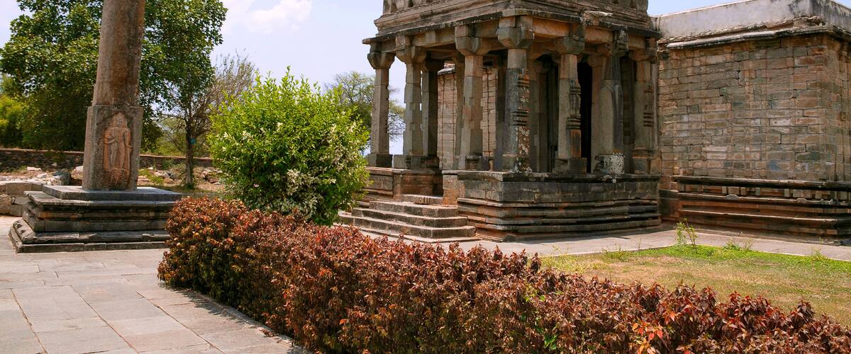 A historical stone structure with columns and a central dome, surrounded by a well-maintained garden with red-leaved bushes.