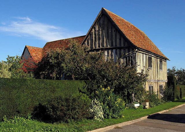 A large timber-framed house with a red-tiled roof, surrounded by lush greenery and a well-maintained hedge.