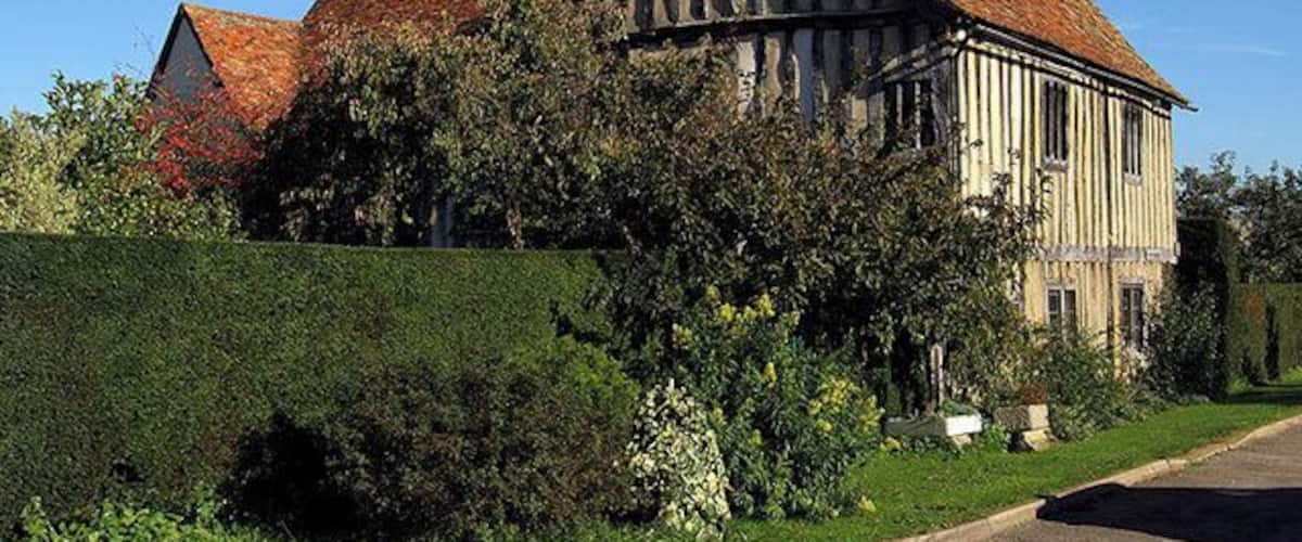 A large timber-framed house with a red-tiled roof, surrounded by lush greenery and a well-maintained hedge.