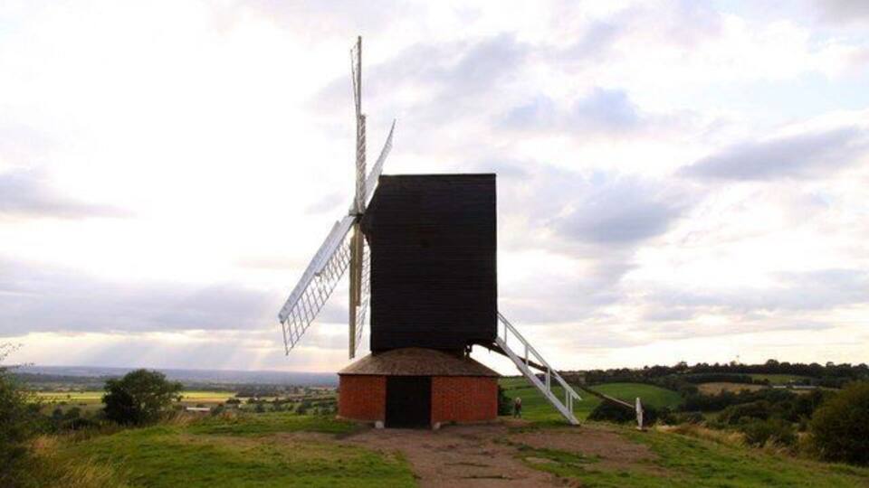 A windmill with a large sail and a brick base, set in a grassy field with a cloudy sky.