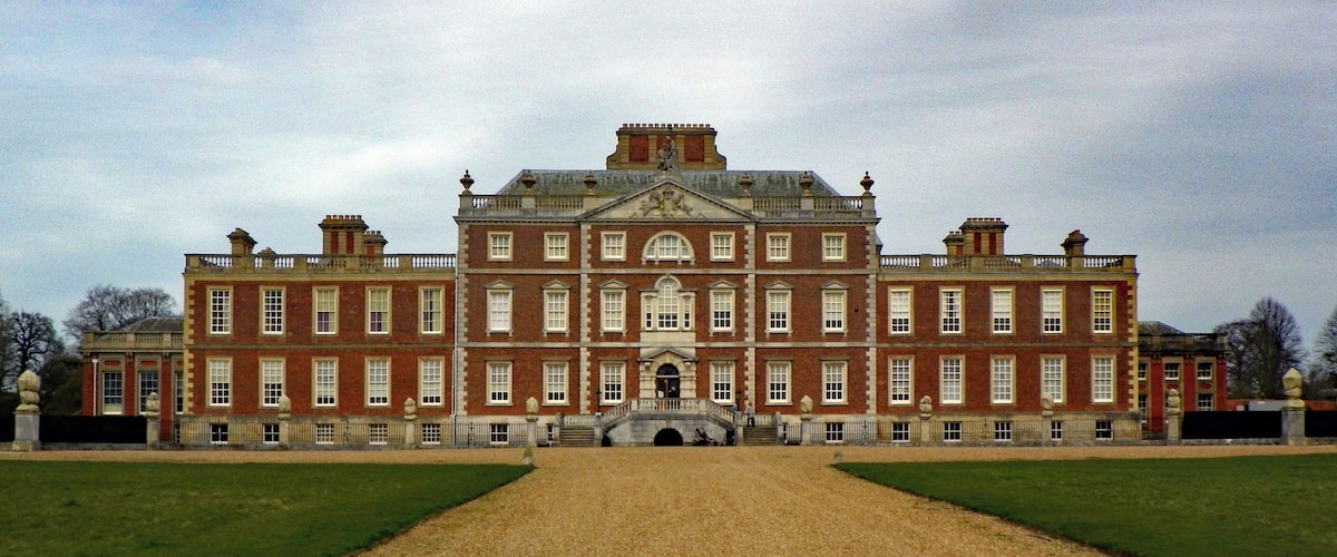 A large, historic red brick building with a symmetrical facade, a central entrance, and a gravel driveway leading to a bridge.