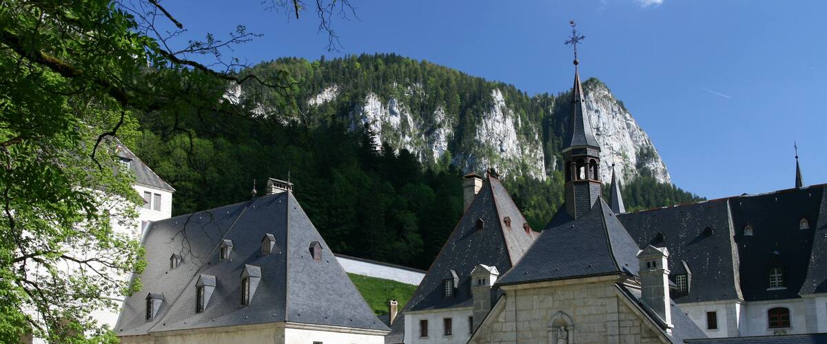 A historic building complex with steeply pitched roofs and a prominent bell tower, set against a backdrop of a forested mountain.