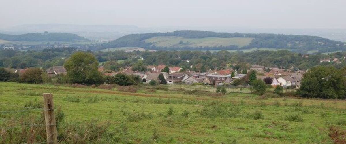 A rural landscape with rolling hills, scattered houses, and a wooden fence in the foreground.
