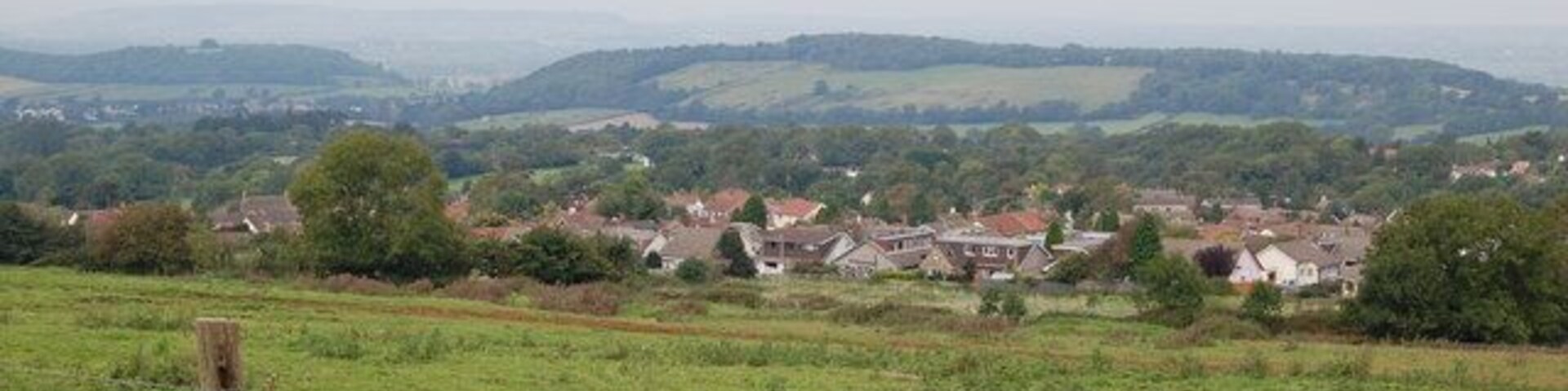 A rural landscape with rolling hills, scattered houses, and a wooden fence in the foreground.