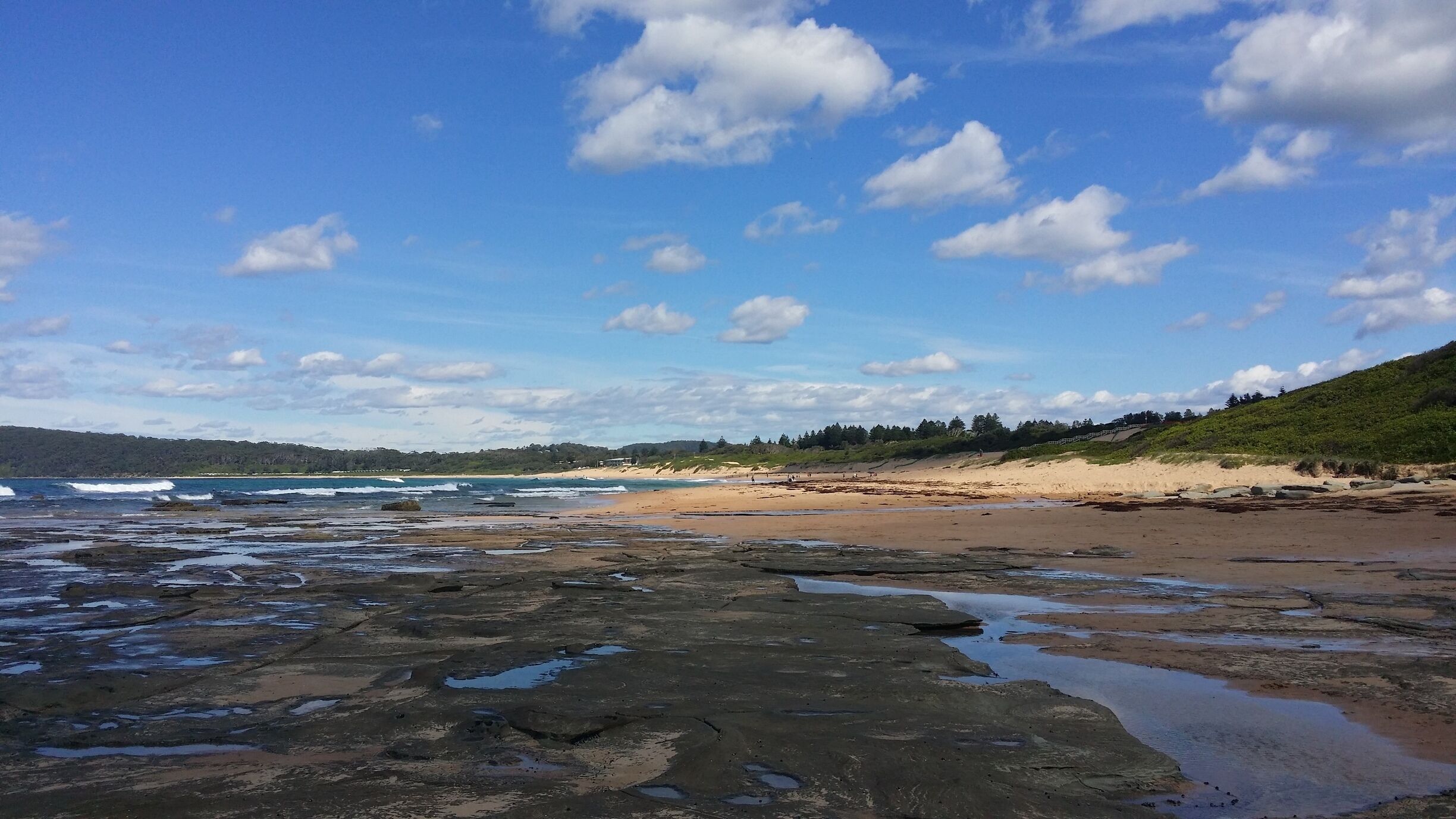 A rocky shoreline with waves crashing onto the beach, a sandy beach, and a forested hill in the background.