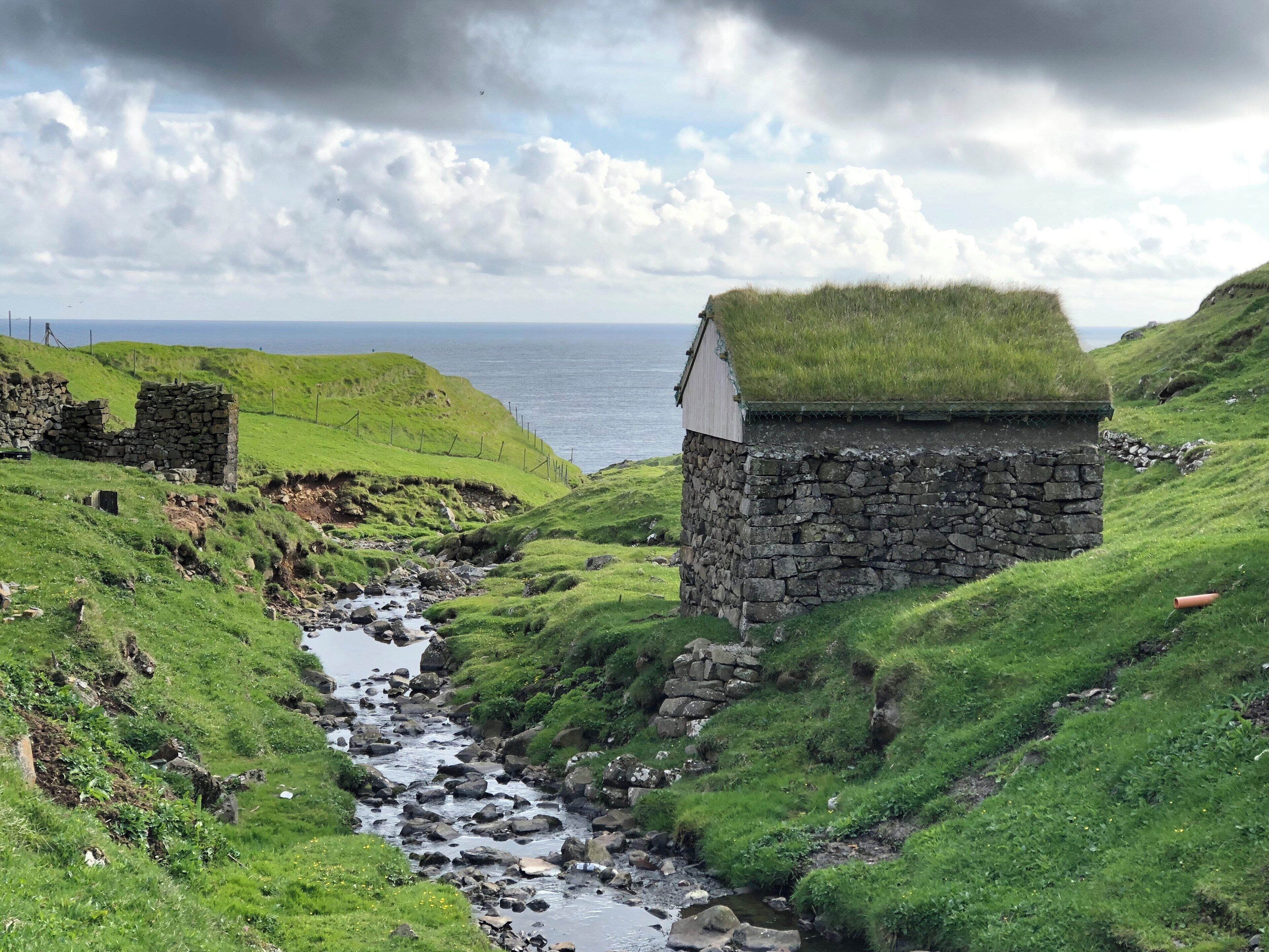 A stone house with a grass roof, a rocky stream, and a grassy hillside.