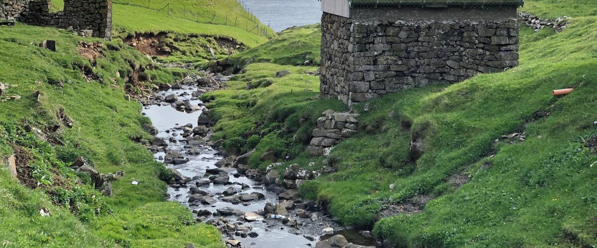 A stone house with a grass roof, a rocky stream, and a grassy hillside.
