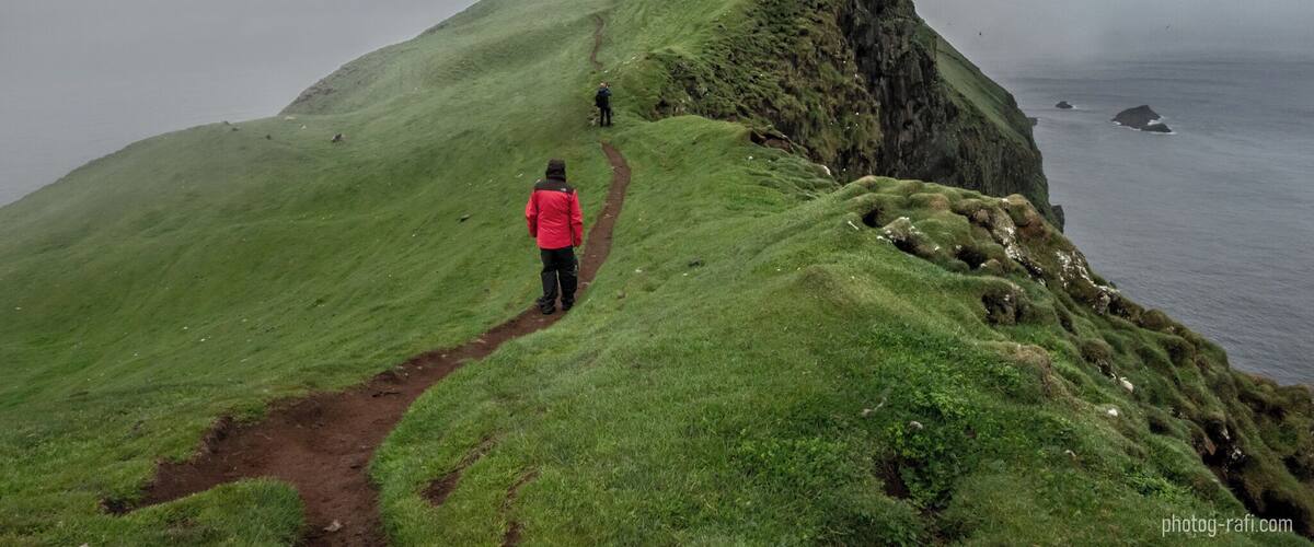 Un randonneur vêtu d’une veste rouge se trouve sur un sentier de montagne recouvert d’herbe, avec une falaise rocheuse et l’océan en arrière-plan.