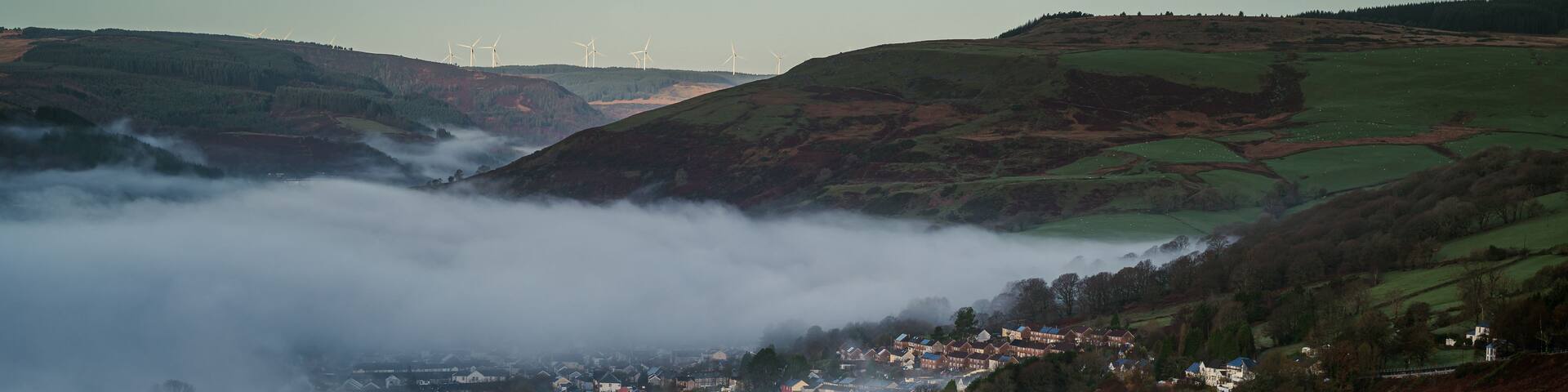 A valley with a town nestled among hills, surrounded by forests and wind turbines in the distance.