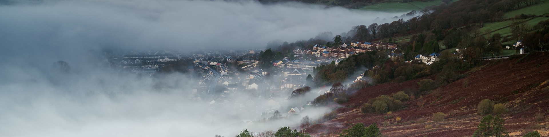 Um vale com uma cidade aninhada entre colinas, cercada por florestas e turbinas eólicas ao longe.