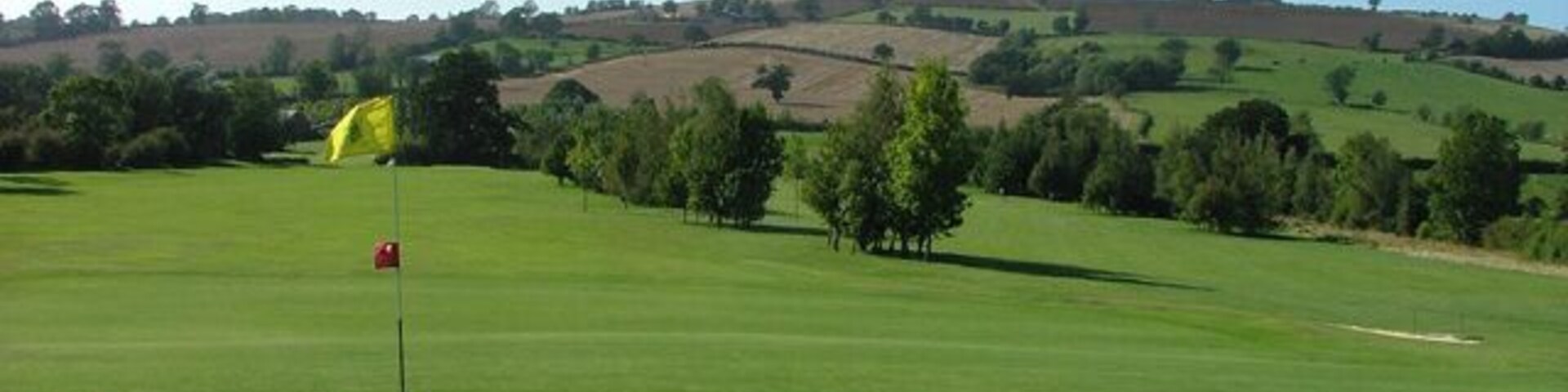 Un campo de golf con un marcador de bandera, árboles y colinas onduladas al fondo.
